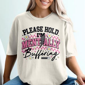 Puede incluir: Camiseta color crema con el texto "PLEASE HOLD I'M MENTALLY Buffering" en negro y rosa. El diseño incluye detalles de margaritas, un rayo y un gráfico de barra de carga. La camiseta es de estilo casual y oversize.