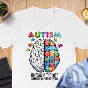 May include: White t-shirt with the word "AUTISM" in colorful letters above a brain graphic. The brain is split, with one half detailed and the other made of puzzle pieces. Text below reads "NO FLAW IN THE CODE DIFFERENT OPERATING SYSTEM."