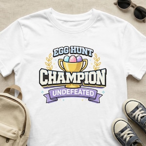 May include: White t-shirt with a colorful graphic. The graphic reads "EGG HUNT CHAMPION UNDEFEATED" with a gold trophy holding pastel colored eggs. The design includes a laurel wreath and a purple banner.