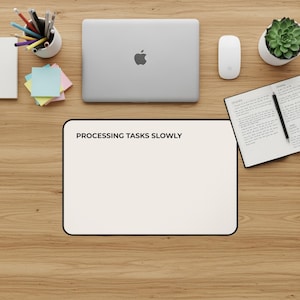 May include: A workspace scene featuring a silver laptop, white mouse, and a notepad with a pen. A white mousepad with the text "PROCESSING TASKS SLOWLY" is in the foreground. A small succulent plant and a pencil holder are also visible.