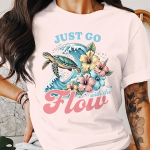 Puede incluir: Camiseta rosa claro con una tortuga nadando en olas azules, rodeada de coloridas flores de hibisco. El texto "JUST GO with the Flow" está impreso en azul y rosa.