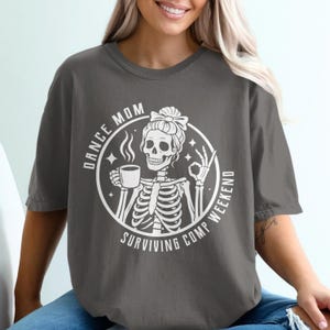 May include: Dark grey t-shirt featuring a white circular graphic. The graphic depicts a skeleton with a bun, holding a coffee cup and giving an okay sign. Text reads "DANCE MOM SURVIVING COMP WEEKEND".