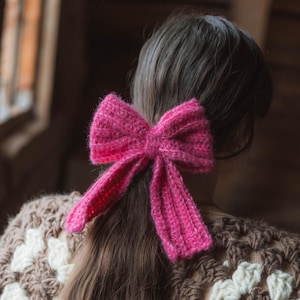 Puede incluir: Un lazo de crochet rosa atado alrededor de una coleta. El lazo está hecho de hilo suave y esponjoso y tiene un aspecto delicado y femenino.