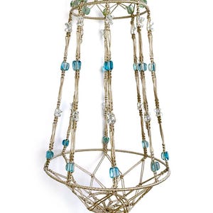 Puede incluir: Un colgador de plantas de macramé beige hecho a mano. Cuenta con cuentas de vidrio azules, verdes y transparentes. El soporte para plantas está suspendido de un anillo de metal y tiene una borla decorativa en la parte inferior.