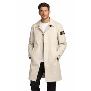 May include: A beige trench coat with a classic design. The coat features a button-up front, a collar, and pockets. A small patch is visible on the left sleeve. The person is wearing a white t-shirt and black pants.