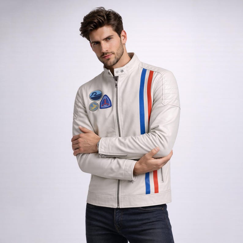 May include: A cream-coloured leather jacket featuring a vertical red and blue stripe on the right side and three patches on the left chest. The jacket has a zip-up front and a stand-up collar. The person in the image is wearing dark blue jeans.