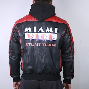 May include: Black leather jacket with red accents on the shoulders and trim. The back features the text "MIAMI VICE STUNT TEAM" in white, blue, and red. The jacket has a black hood and a ribbed hem and cuffs.