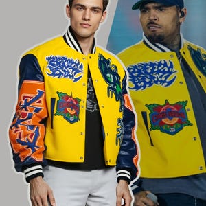 Op de afbeelding: Gele varsity jas met marineblauwe mouwen en oranje accenten. De jas heeft patches met tekst en afbeeldingen, waaronder "CHRIS BROWN 2015". De kraag en manchetten hebben zwart-witte strepen.