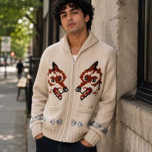 May include: A cream-colored, long-sleeve, zip-up knit cardigan featuring a shawl collar and two front pockets. The sweater displays two symmetrical fox head motifs in brown, black, and red, with a blue paw print pattern along the ribbed hem.