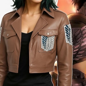 Könnte beinhalten: Braune Lederjacke mit Kragen und zwei Vordertaschen. Die Jacke hat Aufnäher mit einem geflügelten Emblem auf den Ärmeln und der Brust. Die Jacke ist ein Cropped-Style.