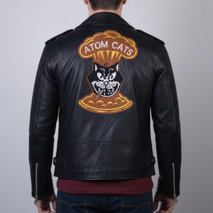 Può includere: Giacca in pelle nera con una grande toppa sul retro raffigurante un gatto dei cartoni animati in una nuvola a fungo atomico con la scritta "ATOM CATS". La giacca ha un classico stile biker con colletto a revers e maniche con zip.