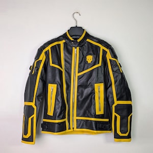 Può includere: Giacca in pelle nera con dettagli gialli e un'emblema a teschio. La giacca presenta una chiusura con zip, diverse tasche e bordi gialli che delineano il design. È appesa a una gruccia nera su sfondo bianco.
