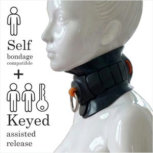 May include: A black bondage collar with a silver metal ring and orange accents. The collar is designed with a padded neck support and a secure closure. The image includes text that says "Self bondage compatible + Keyed assisted release."