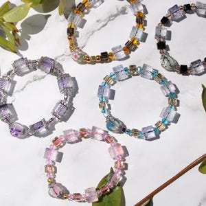 May include: Six bracelets made of clear glass beads with different colored accents. The bracelets are arranged on a white surface with green leaves.