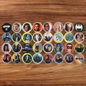 May include: A selection of Batman collectible discs, displaying characters and logos from the franchise. The discs are arranged on a wooden surface, with a variety of colours and designs, including portraits and the bat symbol.