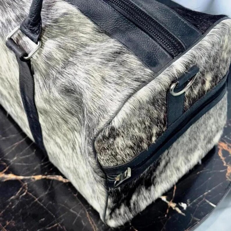May include: A duffel bag with a cowhide exterior in shades of gray and black. The bag features black leather accents, including the handles, straps, and zipper details. The bag is designed for travel and has a rectangular shape.