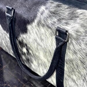 May include: A close-up of a duffel bag with a cowhide pattern in black, white, and gray. The bag has black leather handles and silver-colored metal hardware.