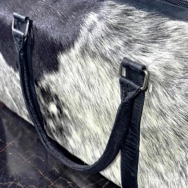 May include: A close-up of a duffel bag with a cowhide pattern in black, white, and gray. The bag has black leather handles and silver-colored metal hardware.