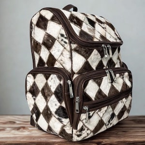 Puede incluir: Mochila de piel de vaca marrón y blanca con un patrón de diamantes. La mochila tiene múltiples compartimentos con cremallera, incluyendo dos bolsillos delanteros y una solapa superior. Las cremalleras son plateadas y los ribetes son marrones. Tiene un asa superior y correas ajustables.