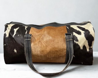 May include: A cowhide duffel bag with a brown center panel and black and white patterned sides. The bag has black leather handles and trim with white stitching. The bag is designed for travel and has a rectangular shape.