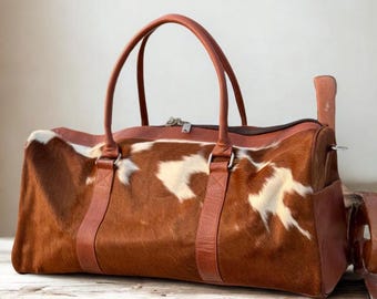 May include: A brown and white cowhide leather duffel bag with brown leather handles and straps. The bag has a zipper closure and a side pocket. The bag is sitting on a wooden surface.