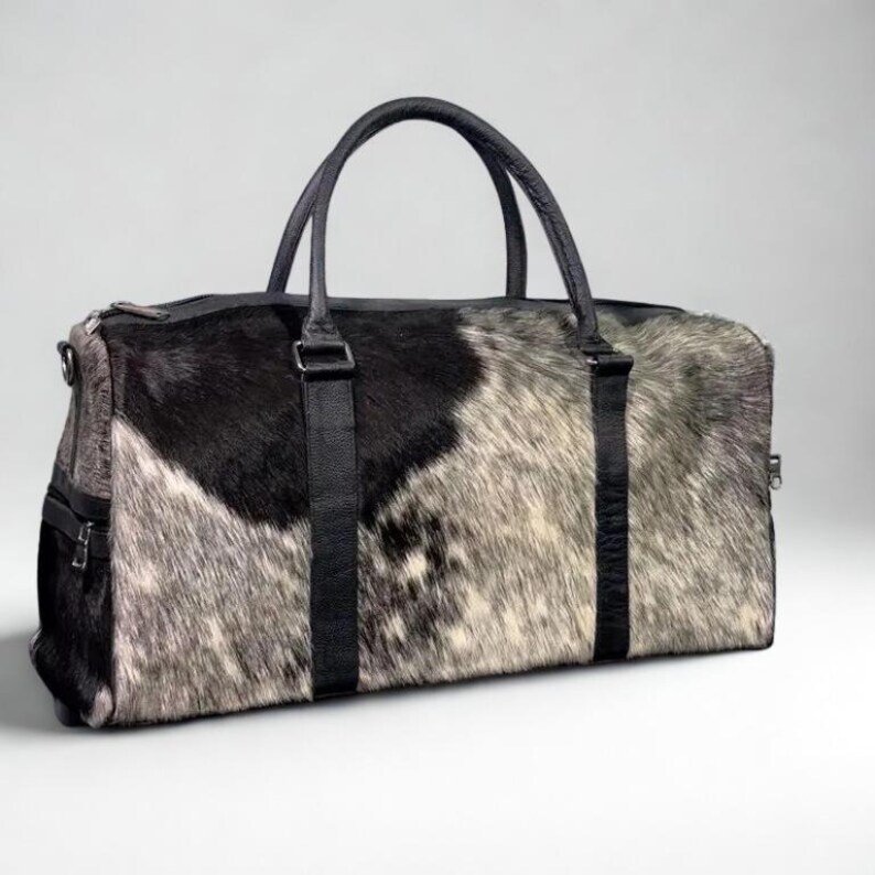 May include: A rectangular duffel bag with a cowhide pattern in black, gray, and white. The bag has black leather handles and straps, with a zipper closure. The bag is designed for travel and has a side pocket.