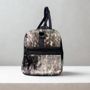 May include: A duffel bag with a cowhide pattern in shades of black, white, and gray. The bag has black leather accents, including the handles, a shoulder strap, and a zippered pocket. The bag is designed for travel.
