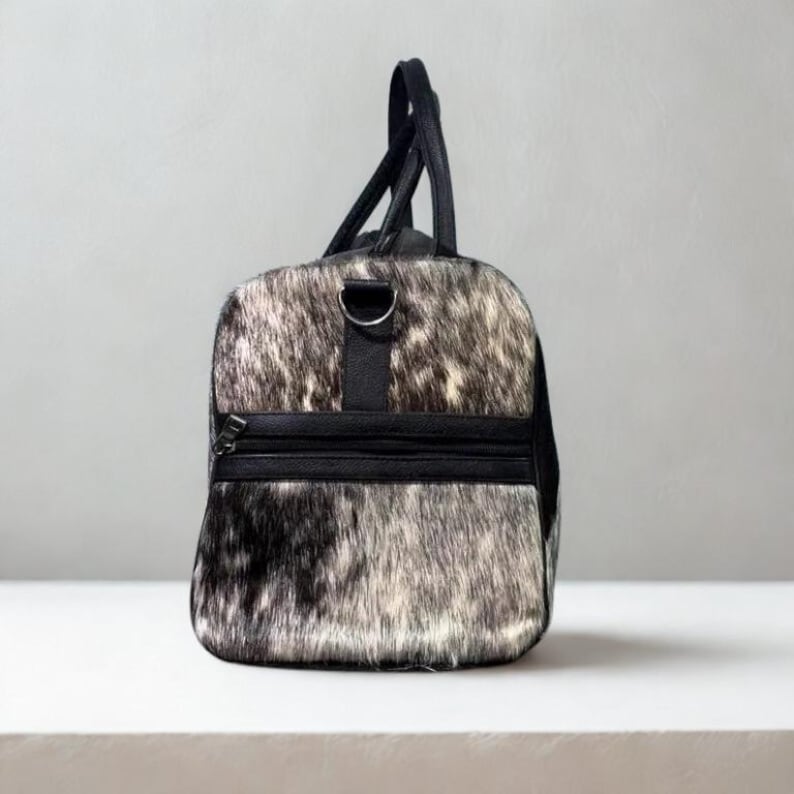 May include: A duffel bag with a cowhide pattern in shades of black, white, and gray. The bag has black leather accents, including the handles, a shoulder strap, and a zippered pocket. The bag is designed for travel.
