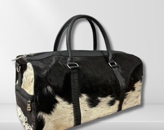 Puede incluir: Una bolsa de viaje de piel de vaca en blanco y negro con asas y ribetes de cuero negro. La bolsa tiene un compartimento principal con cremallera y un bolsillo adicional con cremallera en el lateral. La bolsa es de forma rectangular.