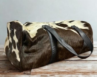 Puede incluir: Una bolsa de lona de piel de vaca con un patrón natural de manchas marrones y blancas. La bolsa tiene asas y ribetes de cuero negro. La bolsa es rectangular y se apoya sobre una superficie de madera.