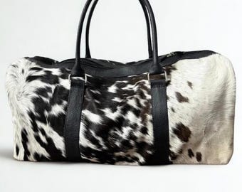 May include: A travel duffel bag featuring a cowhide pattern in black and white. It has black leather handles and straps, and a zip closure. The bag is rectangular in shape and designed for carrying luggage.