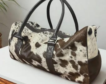 May include: A duffel bag with a cowhide pattern in shades of brown and white. The bag has dark brown leather handles and trim, a zippered top, and a side pocket. The bag is designed for travel or carrying items.