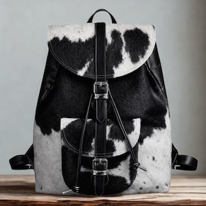 Puede incluir: Mochila de piel de vaca en blanco y negro con cierre de solapa y bolsillo delantero. La mochila tiene correas, hebillas y un asa superior de cuero negro. La bolsa está sobre una superficie de madera.