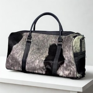 May include: A rectangular duffel bag with a cowhide pattern in black, gray, and white. The bag has black leather handles and straps, with silver-colored metal hardware. The bag is sitting on a white surface.