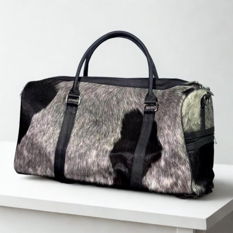 May include: A rectangular duffel bag with a cowhide pattern in black, gray, and white. The bag has black leather handles and straps, with silver-colored metal hardware. The bag is sitting on a white surface.