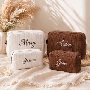 May include: Four personalised cosmetic bags in white and brown waffle weave fabric. The white bags are embroidered with the names "Mary" and "Jassice" in black script. The brown bags are embroidered with the names "Aiden" and "Grace" in white script. Each bag has a zip closure.