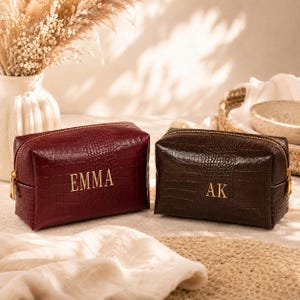 May include: Two rectangular cosmetic bags with a crocodile-embossed texture. The left bag is deep red with the name EMMA in gold lettering. The right bag is dark brown with the initials AK in gold lettering. Both feature gold-toned metal zippers.