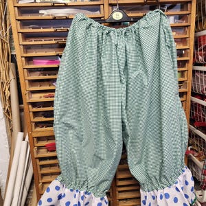 May include: Green and white gingham patterned bloomers with white and blue polka dot ruffled cuffs. The bloomers are hanging on a hanger with the number 14. The background includes a wooden cabinet with drawers and spools of thread.