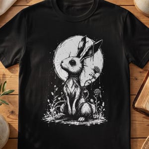 Peut inclure: Un t-shirt noir avec une illustration graphique blanche représentant un lapin assis devant une grande lune circulaire. Le lapin est entouré de vignes de roses épineuses et de petites fleurs, dans un style détaillé de croquis en noir et blanc.