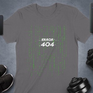 Może przedstawiać: Ciemnoszary t-shirt z krótkim rękawem z centralną grafiką przedstawiającą biały napis ERROR 404 nałożony na pionowy wzór zielonego cyfrowego kodu binarnego. Koszulka jest otoczona smartfonem, smartwatchem, głośnikiem i hantlami.