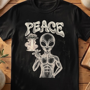 May include: A black t-shirt featuring a white graphic illustration of an alien holding a cigar. Above the alien, the word PEACE is printed in a stylised font. To the left of the alien, a flying saucer emits a beam of light onto a cloud.