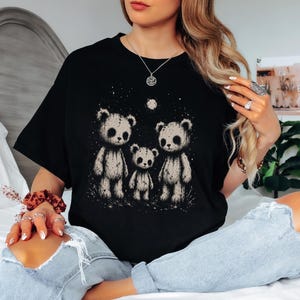 Può includere: Una t-shirt nera con una stampa grafica di tre orsacchiotti beige in piedi insieme sotto una piccola luna e stelle. Gli orsi hanno un aspetto usurato e strutturato con occhi scuri e cavi. La maglietta ha un girocollo.