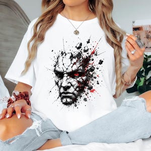 Può includere: Una t-shirt bianca a maniche corte con una stampa grafica di un volto incrinato simile alla pietra con un occhio rosso brillante, circondato da schizzi di inchiostro nero e rosso centrati sul petto del capo.