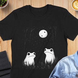 May include: Black t-shirt featuring a whimsical illustration of two white frogs gazing at a full moon in a starry night sky. The design includes stylized trees and grass, creating a serene, nature-inspired scene.
