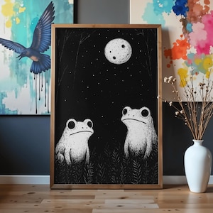 May include: A framed black and white illustration depicts two white frogs gazing at a full moon in a starry night sky. The artwork is set in a dark forest with tall grass, framed in a gold-colored wooden frame.