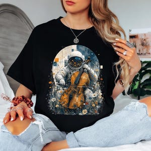 Może przedstawiać: Czarny t-shirt z nadrukiem graficznym przedstawiającym astronautę w białym skafandrze kosmicznym grającego na złotobrązowej wiolonczeli. Astronauta jest otoczony polem małych białych i pomarańczowych kwiatów na okrągłym, teksturowanym tle.