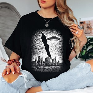 May include: A black t-shirt featuring a detailed white graphic print of a flying saucer hovering above a stone circle formation. The design includes a dark, vertical cloud-like shape extending from the sky towards the ancient stone structures.