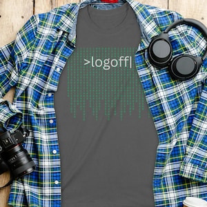 Peut inclure: Un t-shirt gris foncé avec le texte ">logoff|" et un motif vert de style matrice. Une chemise à carreaux bleue, verte et blanche est drapée sur le t-shirt. Un casque noir et un appareil photo sont également visibles sur une surface en bois.