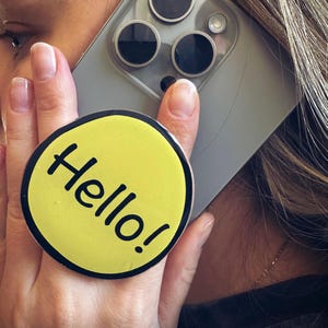 May include: A yellow phone grip with the word "Hello!" in black, held on the back of a silver phone. The grip has a black border. The phone is held up to the ear.