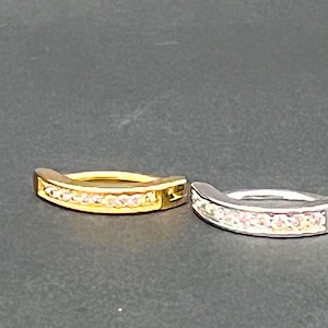 May include: Two curved rings, one gold-toned and one silver-toned, each adorned with a row of small, sparkling gemstones. The rings are set against a dark background, highlighting their metallic sheen and the brilliance of the stones.